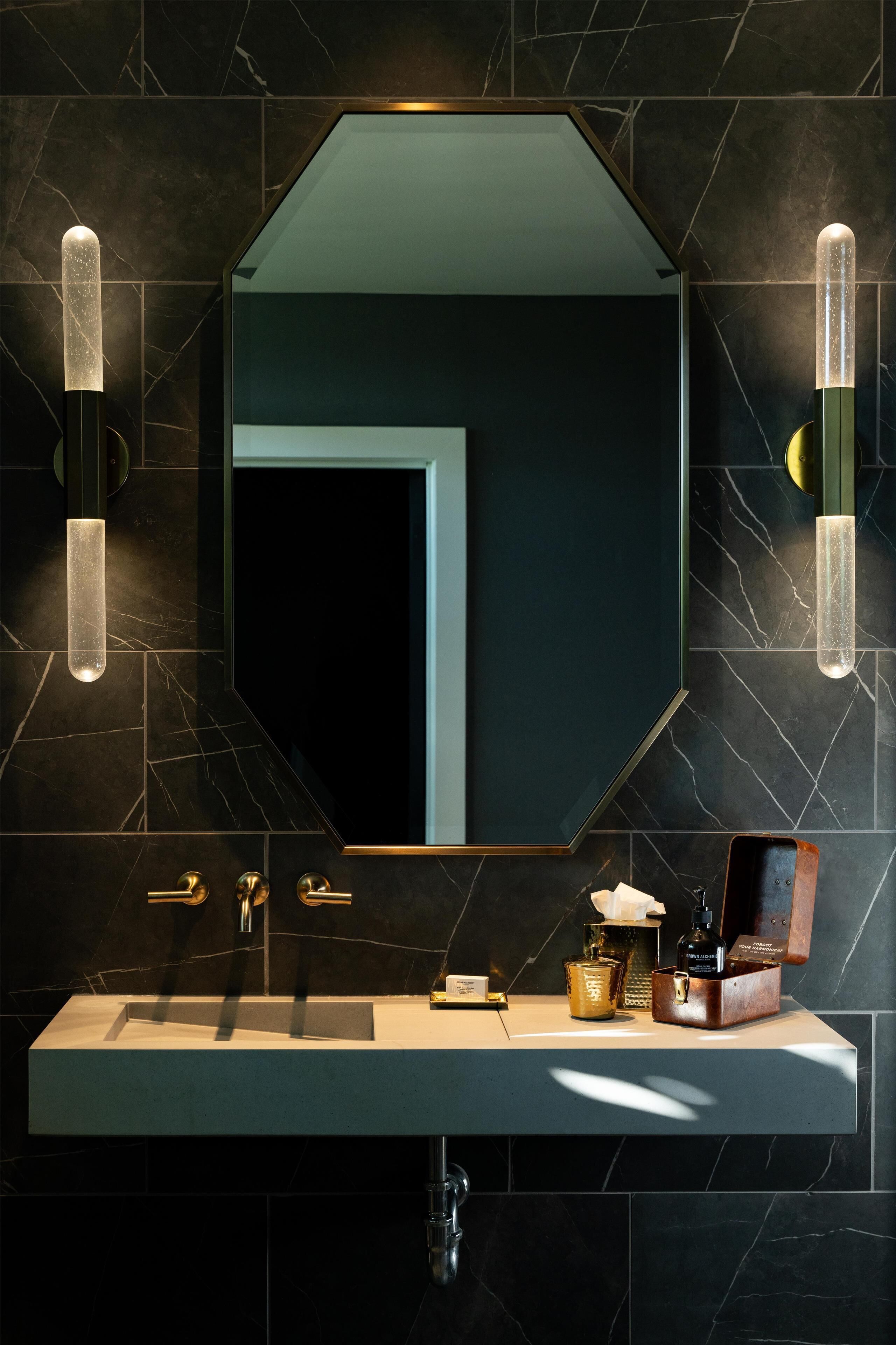 A modern bathroom with a large mirror, marble tiles, a sink with gold fixtures, and a bottle of perfume.