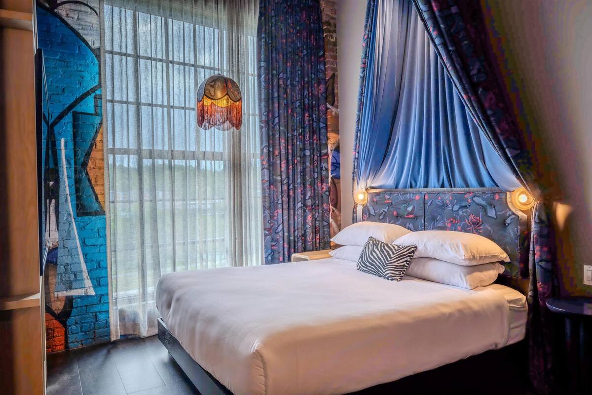 A bedroom with a bed, curtains, a hanging light fixture, and a wall with a blue and red design.