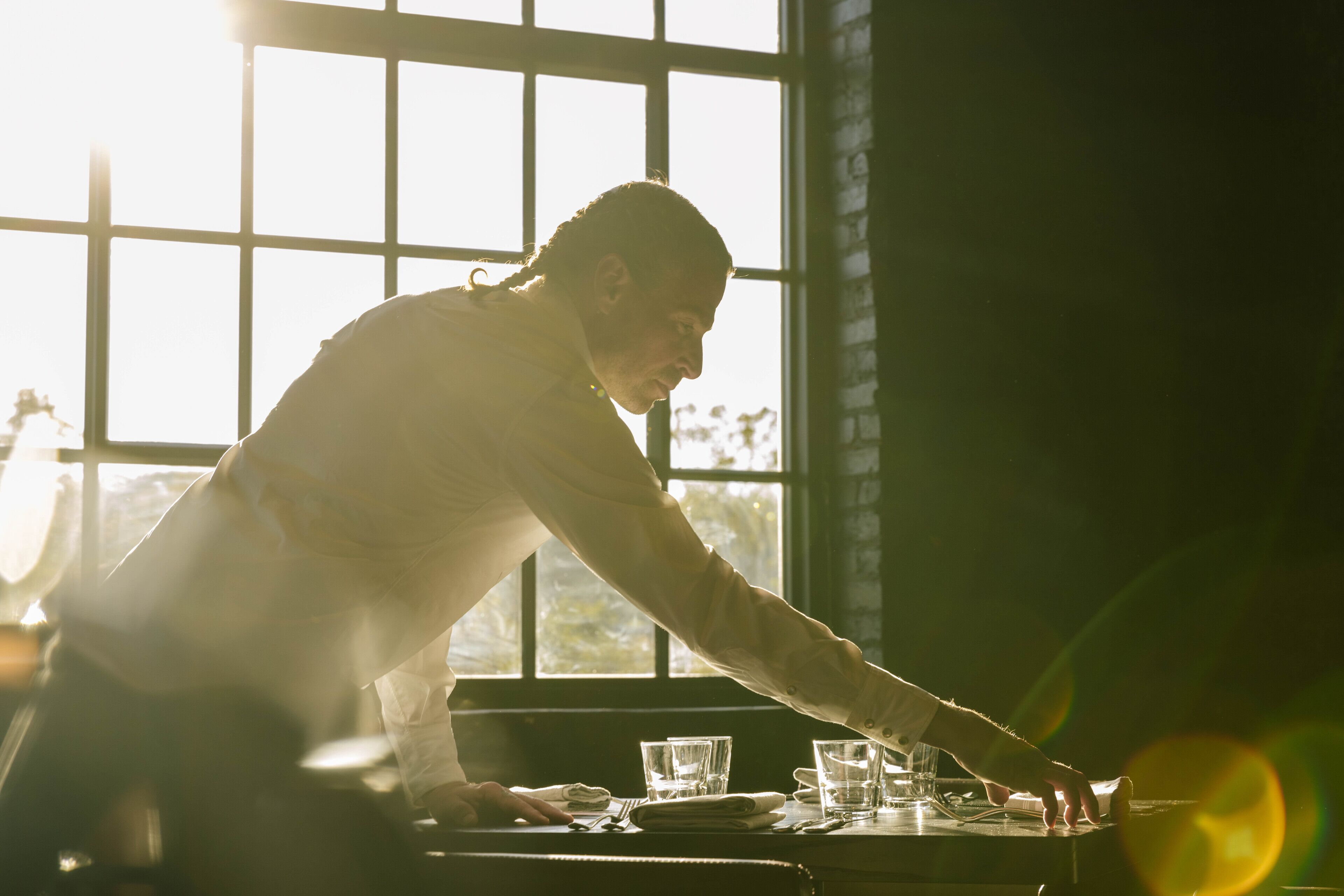 A person setting a table with glasses and plates, in a room with large windows and a brick wall.