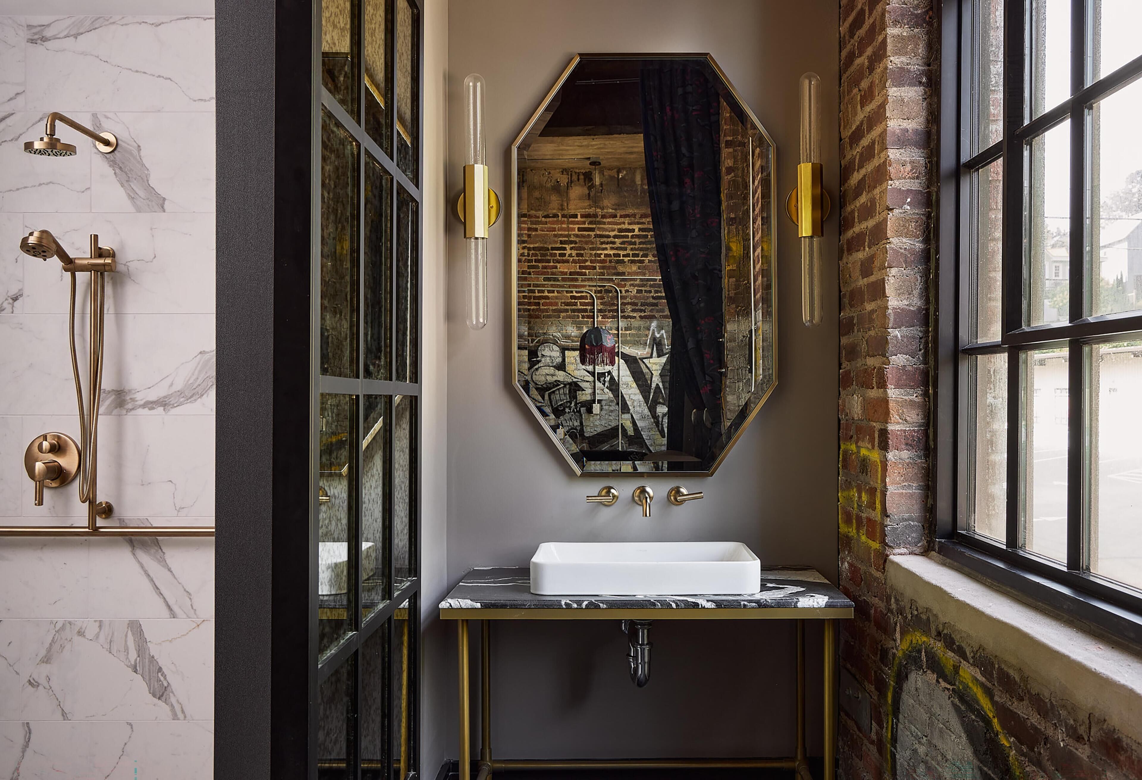 A bathroom with a marble wall, a large mirror, a white sink, and a shower with a gold faucet.