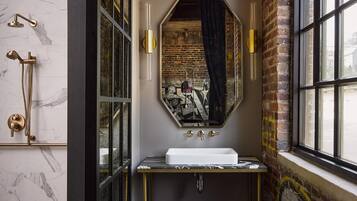A bathroom with a marble wall, a large mirror, a white sink, and a shower with a gold faucet.
