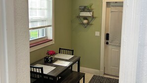 Uma sala de jantar com uma mesa posta para dois, uma janela com persianas, uma porta e uma estante fixada na parede com objetos decorativos.