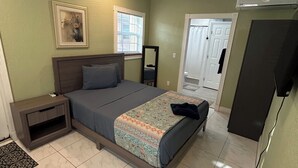 Quarto com cama, mesa de cabeceira, ventoinha no teto e uma porta que dá para outro cômodo.