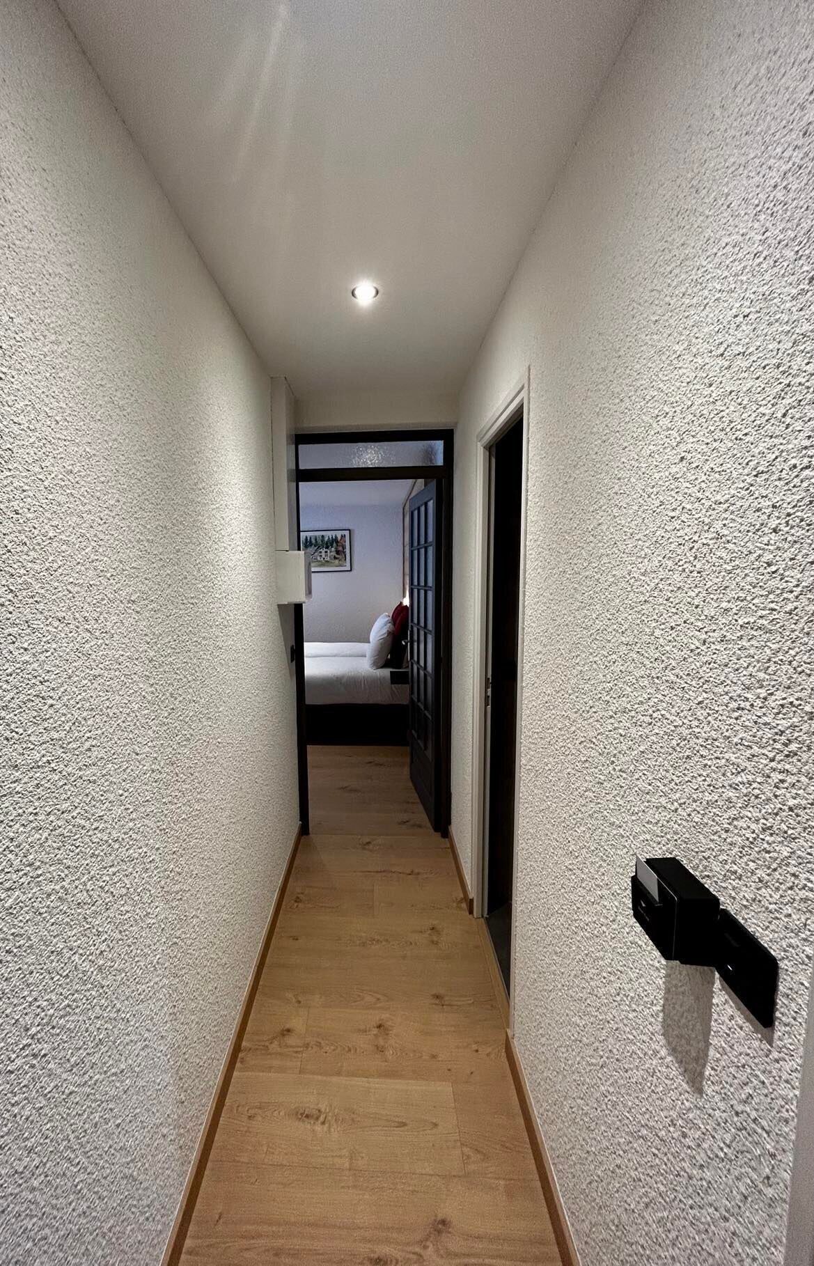 Un pasillo estrecho con paredes texturizadas y piso de madera que conduce a un dormitorio con cama y ventana.
