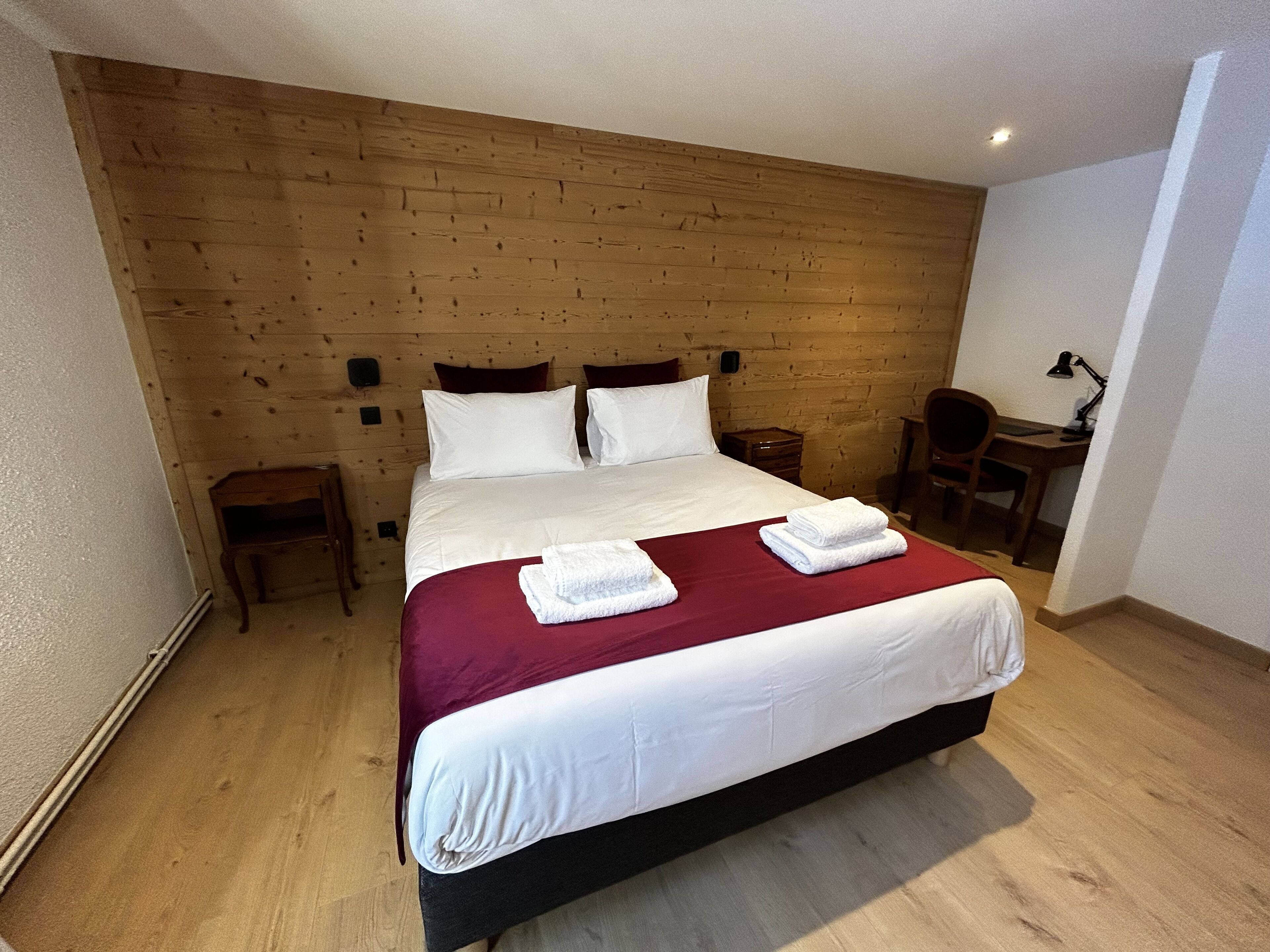 A neatly made bed with white linens and two folded towels, a wooden headboard, a small wooden nightstand, a desk with a chair, and a lamp.