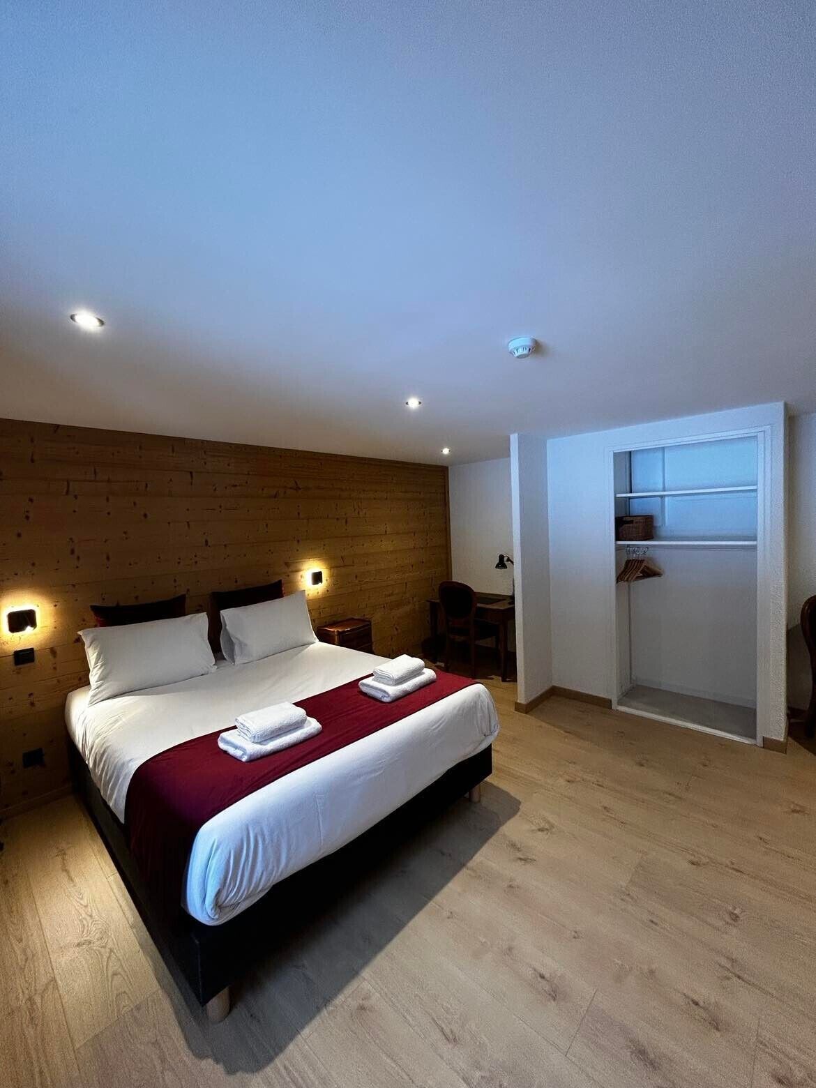 Een moderne slaapkamer met een houten hoofdbord, een groot bed met witte lakens, een rode plaid en een klein bureau met een stoel.