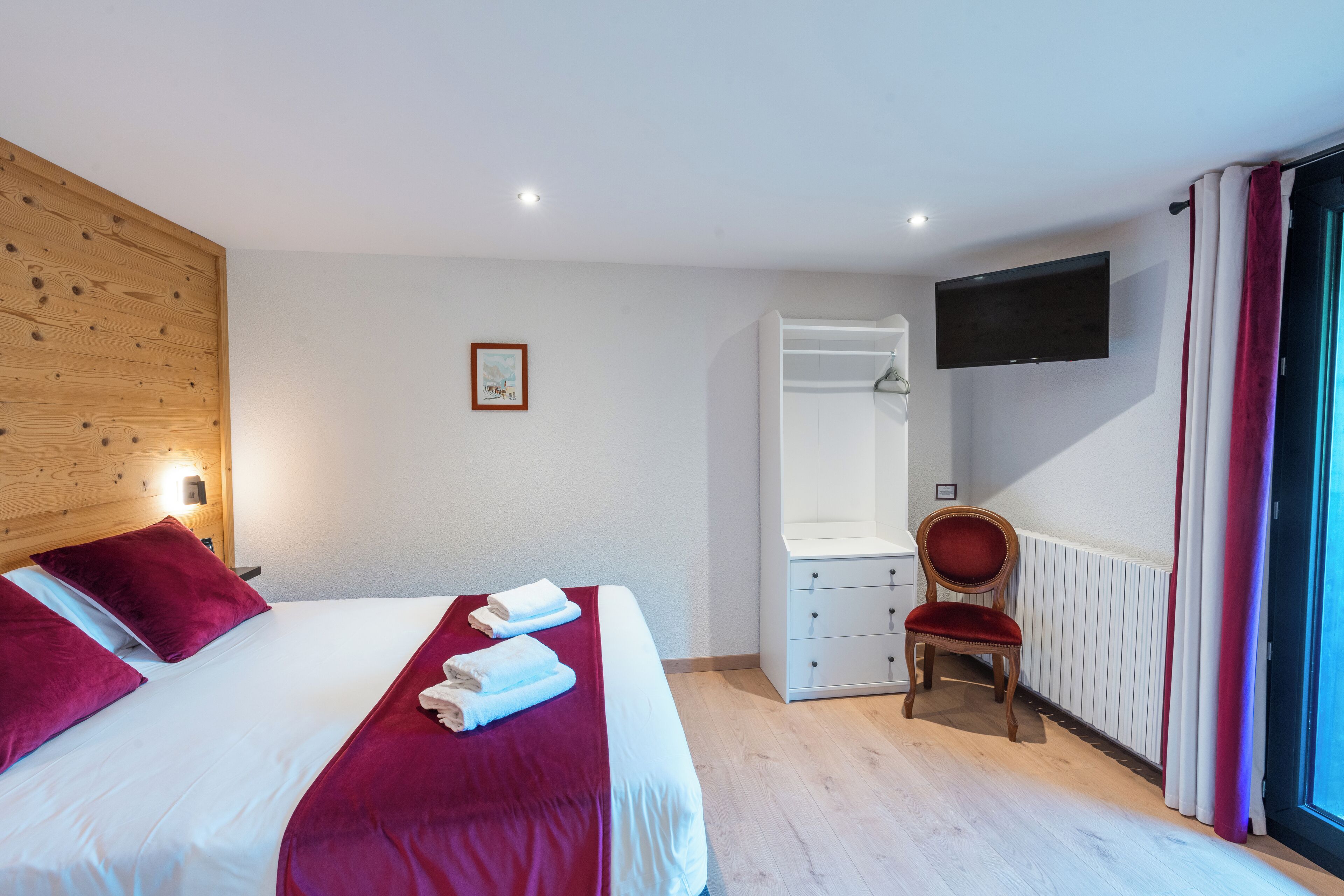 Camera d'albergo con una testiera in legno, un letto grande con biancheria rossa, una sedia, una cassettiera, un televisore montato a parete e una finestra con tende.