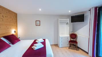 Camera d'albergo con una testiera in legno, un letto grande con biancheria rossa, una sedia, una cassettiera, un televisore montato a parete e una finestra con tende.