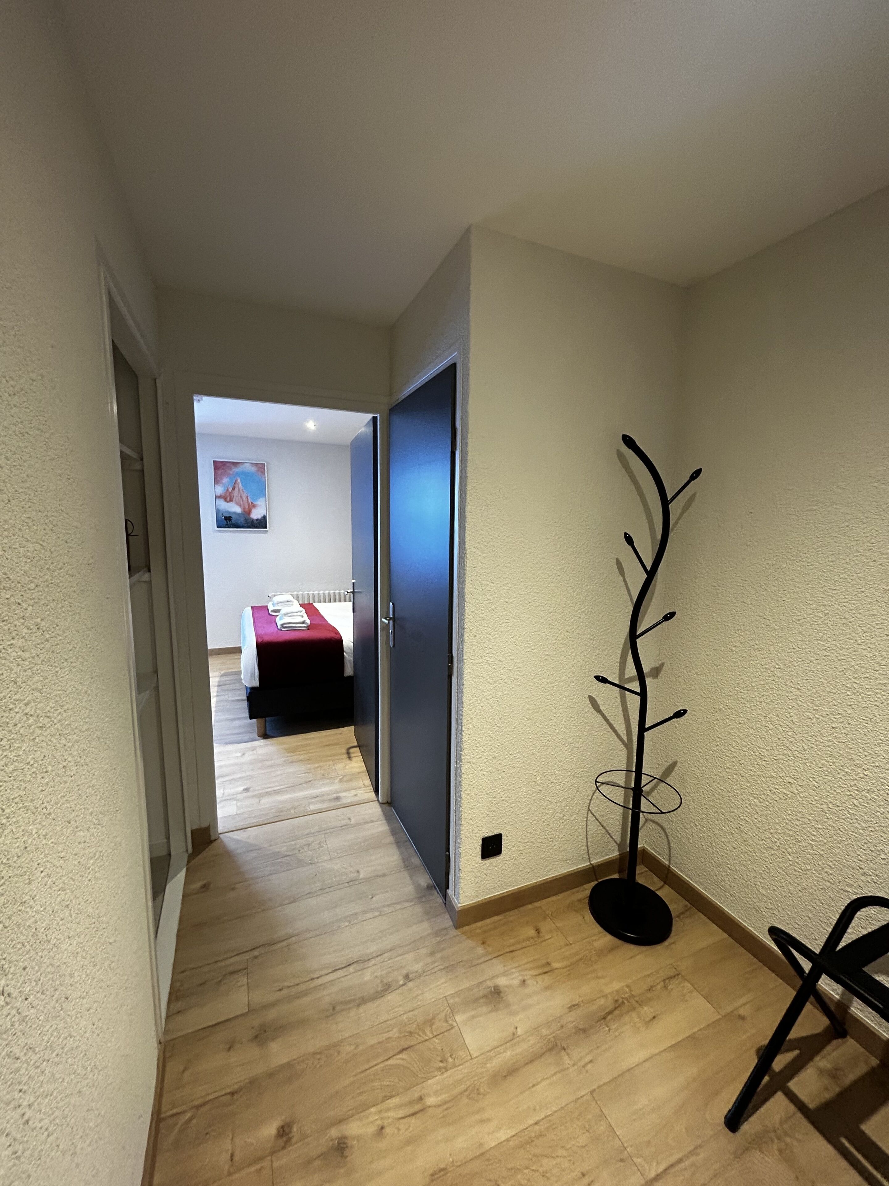 A hallway with wooden flooring leading to a bedroom with a red bedspread. A coat rack is visible on the right wall.