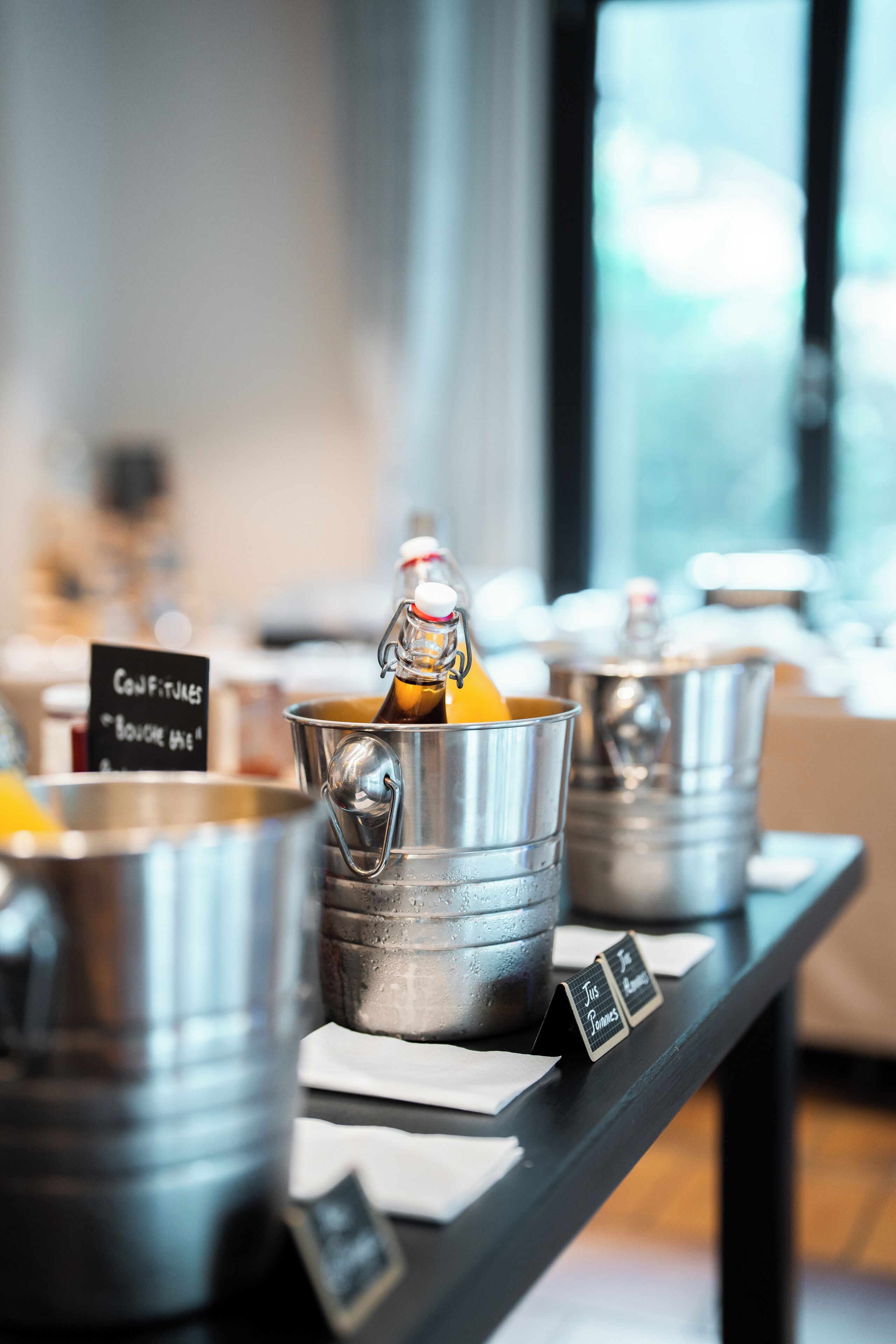 Tavolo del buffet con diversi cibi e condimenti in secchi metallici, una lavagna con il menu e un cartello che indica 'Confitures' e 'Bouquet'.