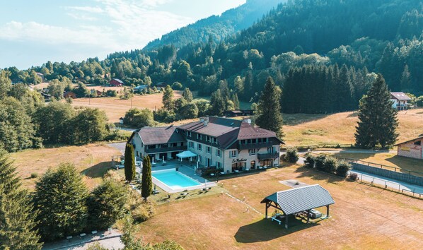 Une grande maison avec une piscine, entourée d’arbres et située dans un paysage vallonné.