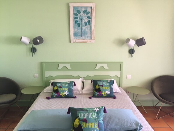 Une chambre avec une tête de lit verte, deux oreillers blancs, deux coussins aux motifs tropicaux évoquant l’été, un tableau encadré représentant un palmier et deux lampes fixées au mur.