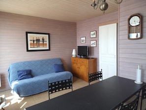 A room with a blue patterned sofa, a wooden cabinet with a television, a dining table with chairs, and a wall clock.