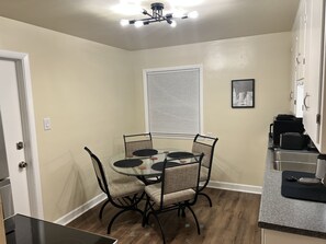 Une cuisine compacte avec un coin repas, comprenant une table ronde en verre avec des chaises, un four encastré et une fenêtre avec des stores.