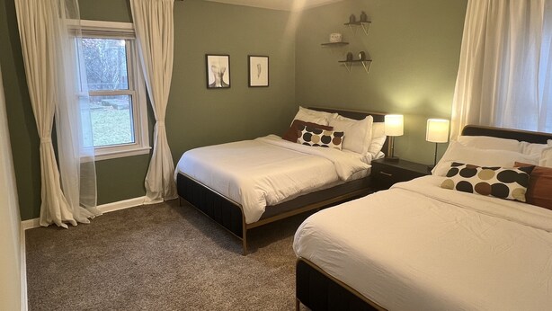 A bedroom with two beds, a window with curtains, a wall-mounted shelf with decorative items, and a ceiling light fixture. - EKO Stay - Your Home Away From Home (Wauwatosa)