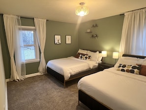 Une chambre avec deux lits, une fenêtre avec des rideaux, une étagère fixée au mur ornée d’objets décoratifs et un luminaire au plafond.