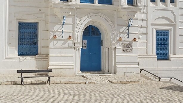 A white building with blue doors and windows, a flag, and a cobblestone courtyard. - Medina town house (Sousse)