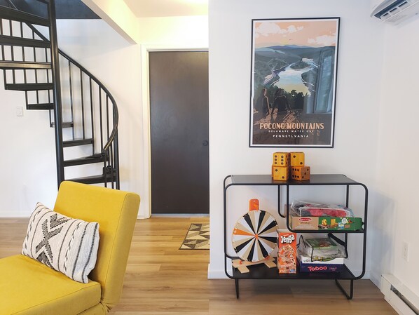 A living room with a yellow sofa, a black metal shelf with board games, and a framed picture of Pocono Mountains.