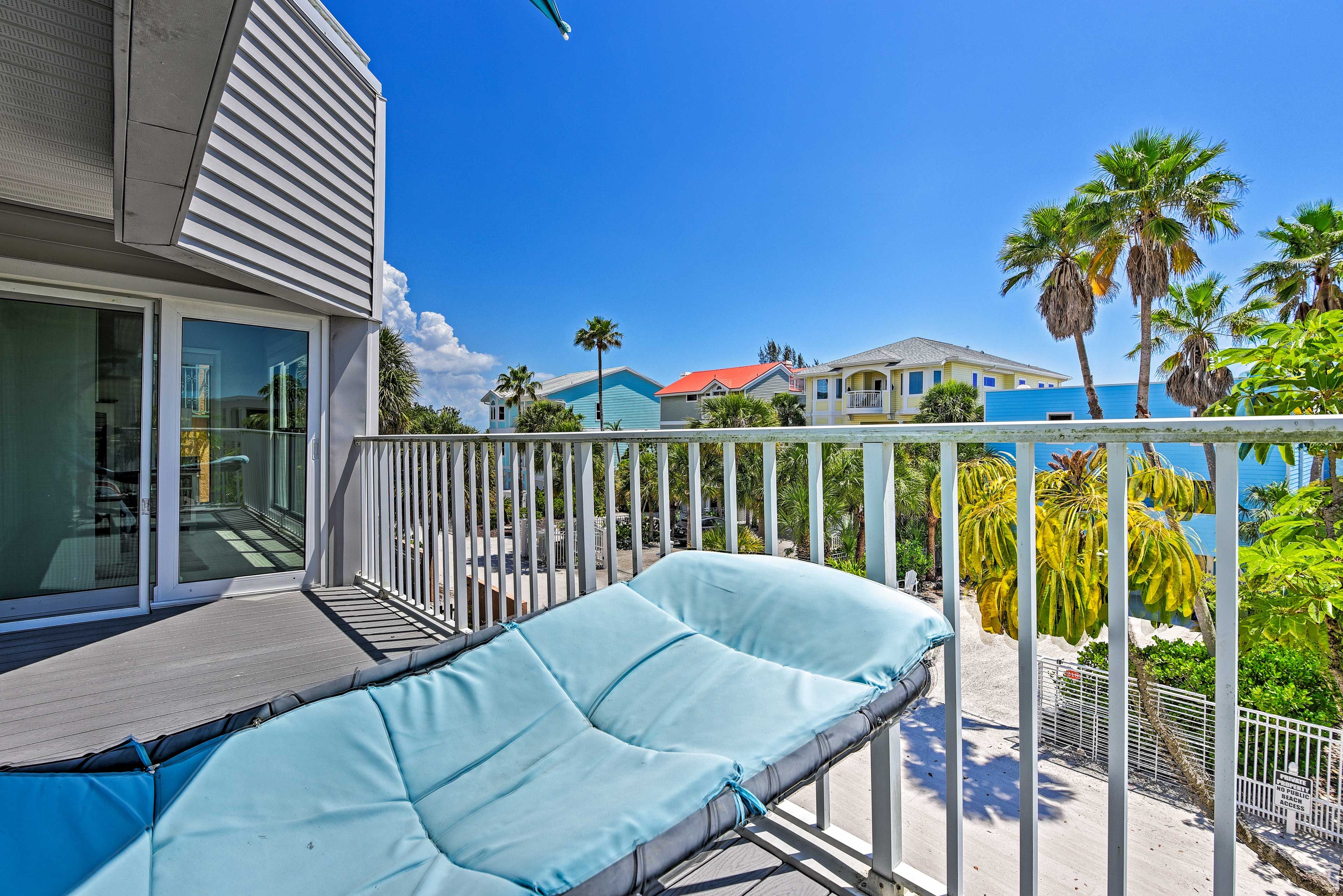 A balcony with a cushioned seating area, a pool, and a view of neighboring houses and palm trees.