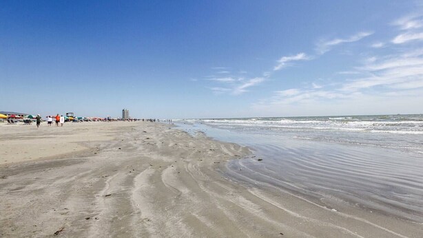 Ein weiter Sandstrand mit sanften Wellen, blauem Himmel und Gebäuden in der Ferne.