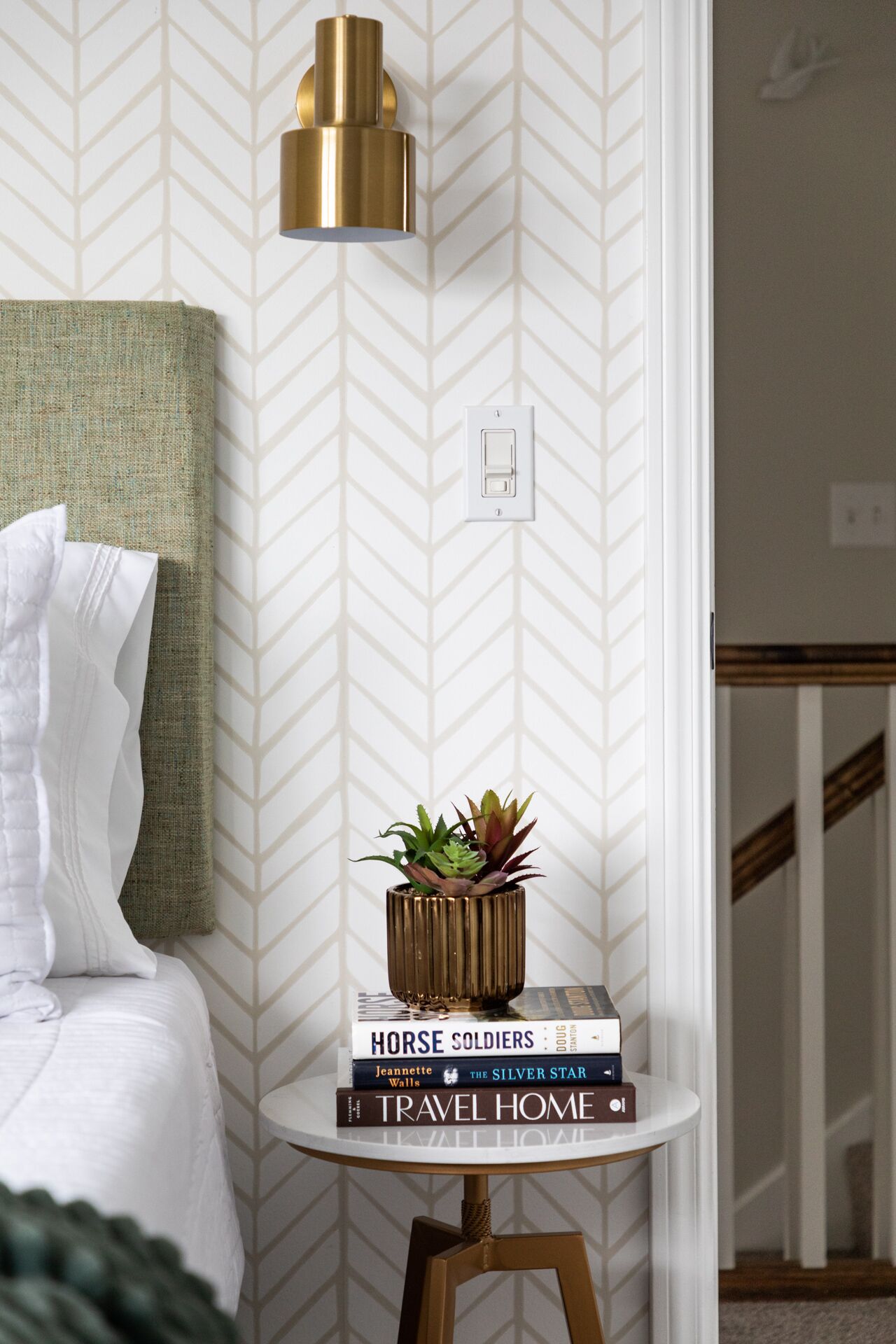 A neatly arranged bedroom with a bed, a bedside table with books, a small potted plant, and a wall-mounted light fixture.