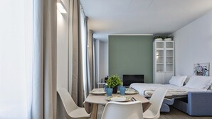 Ett modernt, väl upplyst rum med ett matbord för två personer, en säng med en grå sänggavel och ett vitt skåp med hyllor.