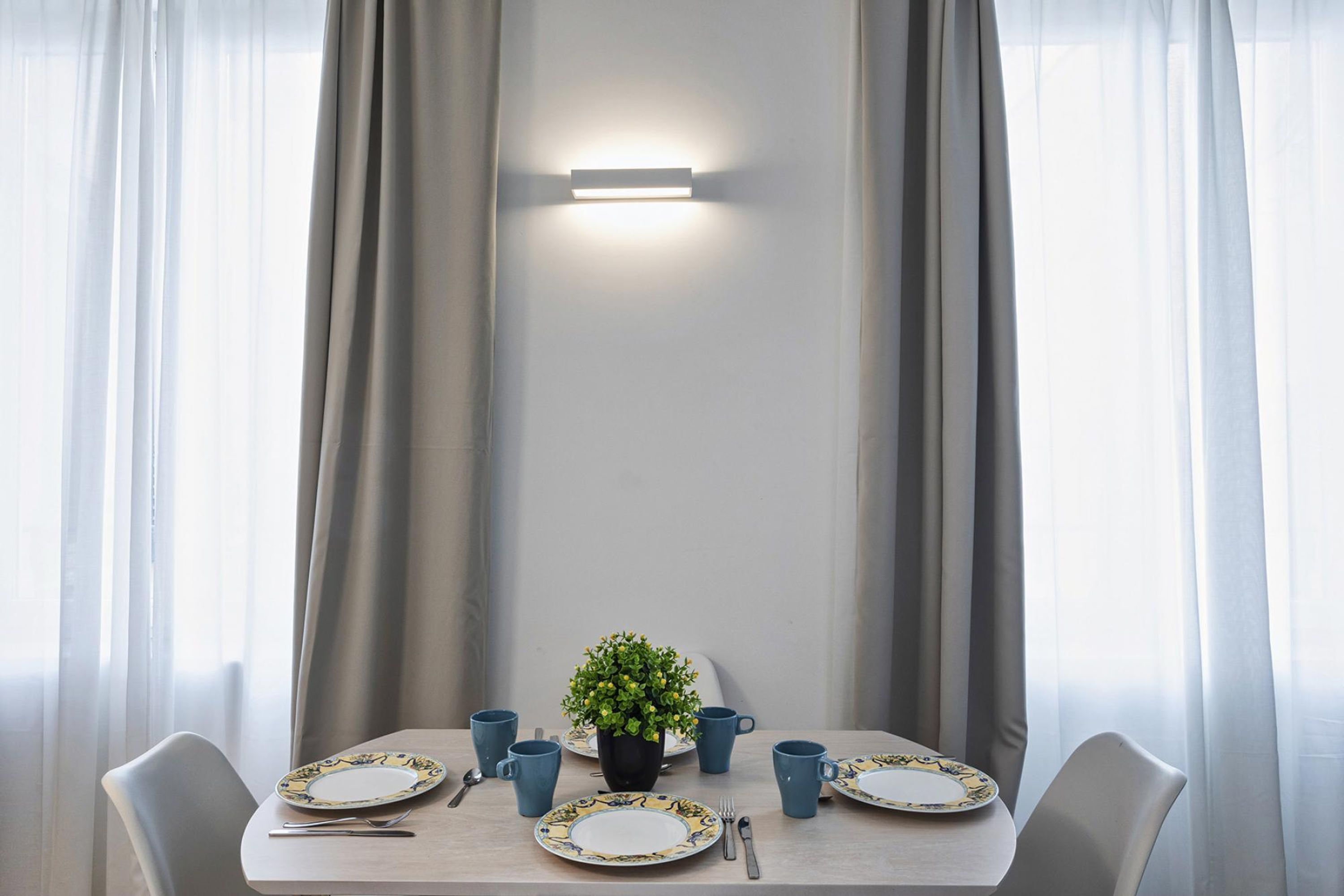 Et spisebord dækket med tallerkener, kopper og bestik, med gardiner og en vægmonteret lampe på hver side.