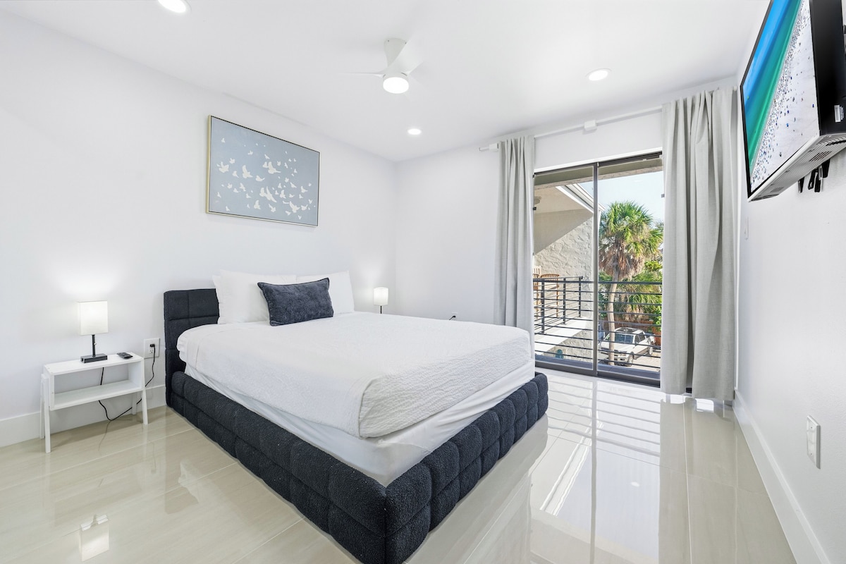A modern bedroom with a large bed, a nightstand, a flat-screen TV, and a balcony with a view of palm trees and a car.