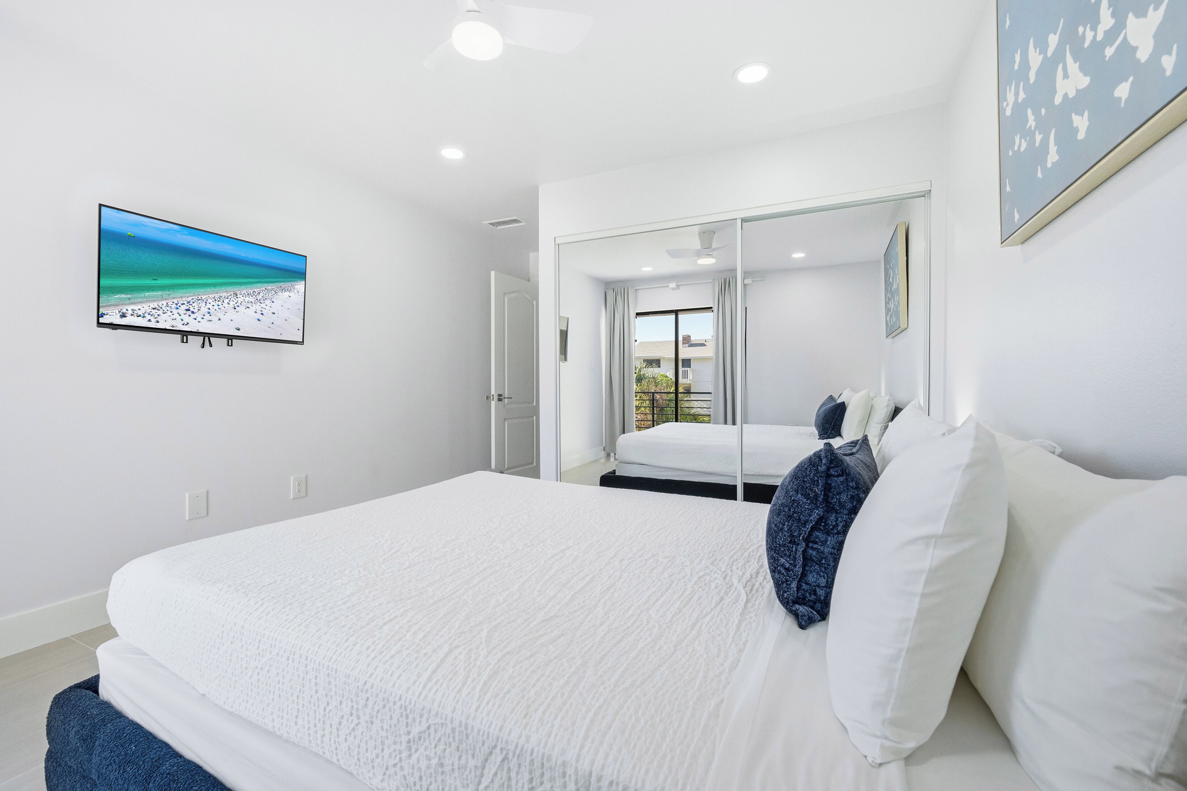 A modern hotel room with a large bed, a television displaying a beach scene, a mirror, and a view of the outdoors.