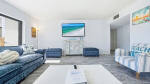 A living room with a sofa, ottoman, and armchair, a flat-screen TV mounted on the wall, and a beach-themed painting. - Just Beachy by Beachside Management (Siesta Key)