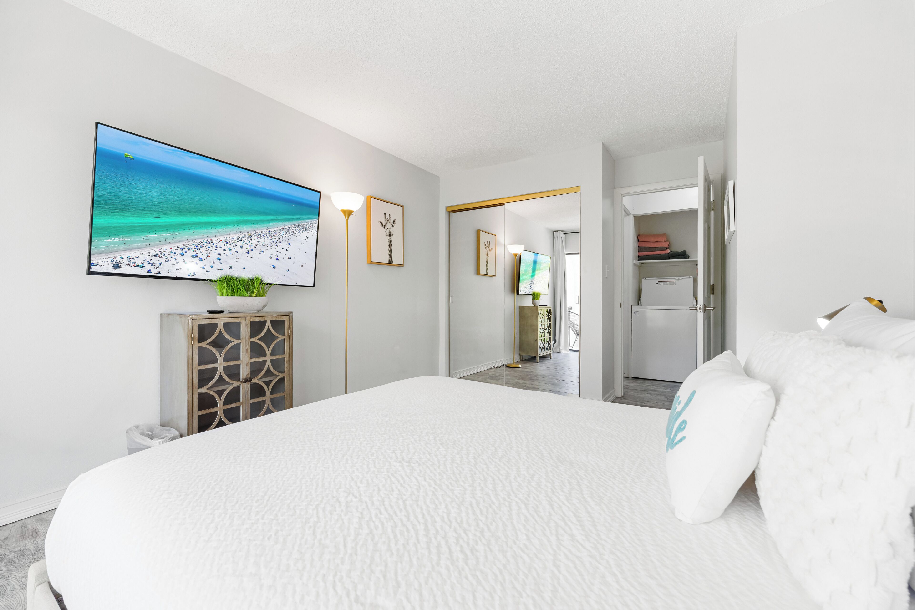 A bedroom with a large bed, a flat-screen TV on the wall, a plant on a cabinet, and a closet.