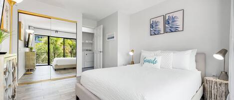 A modern bedroom with a large bed, a bedside table, a nightstand, and a view of the kitchen and balcony.