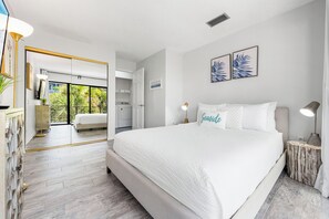 A modern bedroom with a large bed, a bedside table, a nightstand, and a view of the kitchen and balcony.