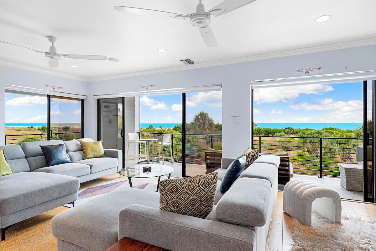 A spacious living area with a grey sofa set, a glass coffee table, and a view of the ocean through large sliding doors.