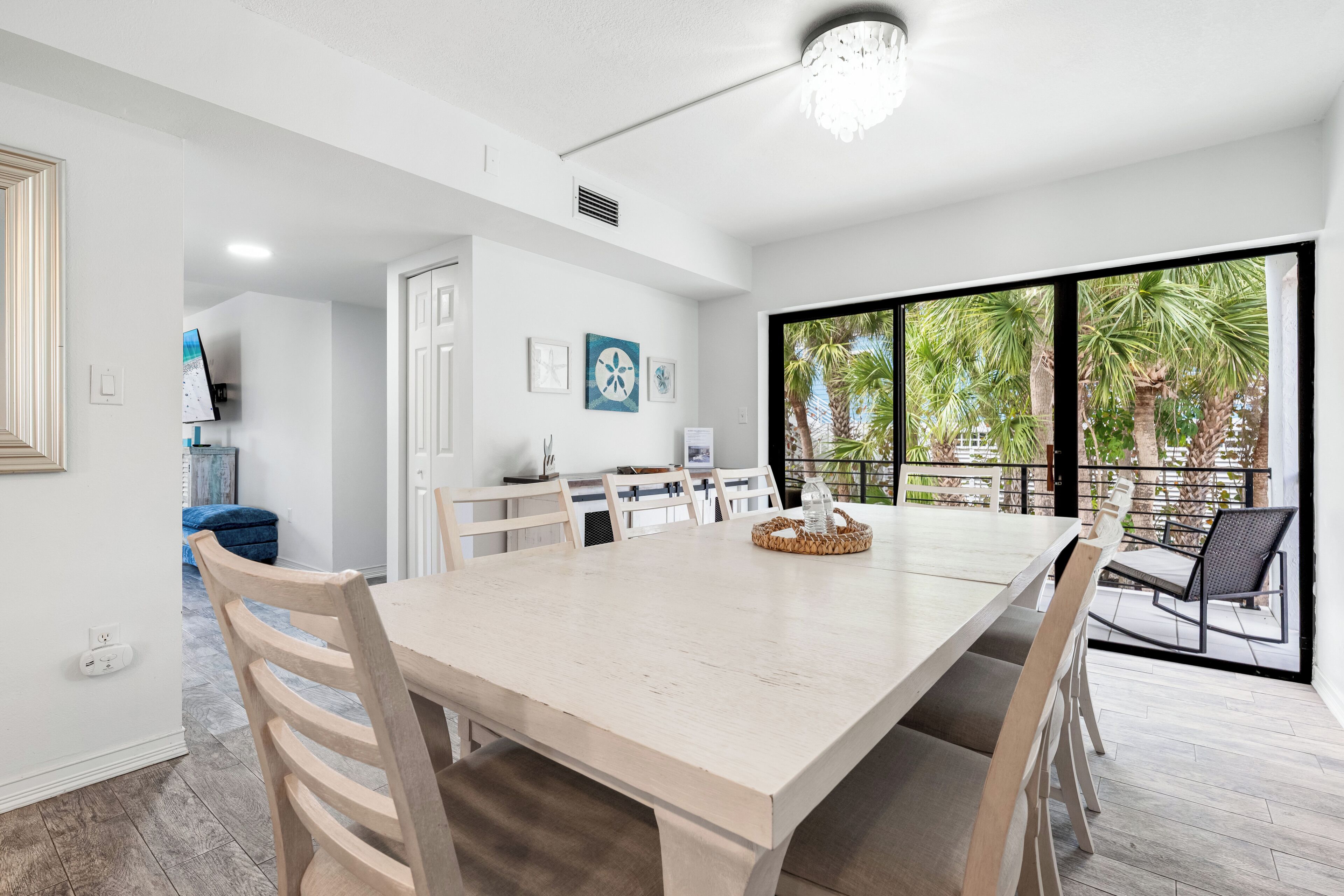 A dining area with a large table, chairs, and a view of palm trees through large windows.