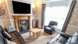 A living room with a fireplace, leather sofas, a TV, and a view of the outdoors. - Coastguard Cottages (Hull)