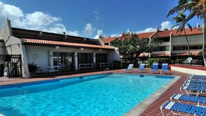 Uma piscina com água azul, rodeada por espreguiçadeiras e um edifício com telhado de telhas vermelhas.