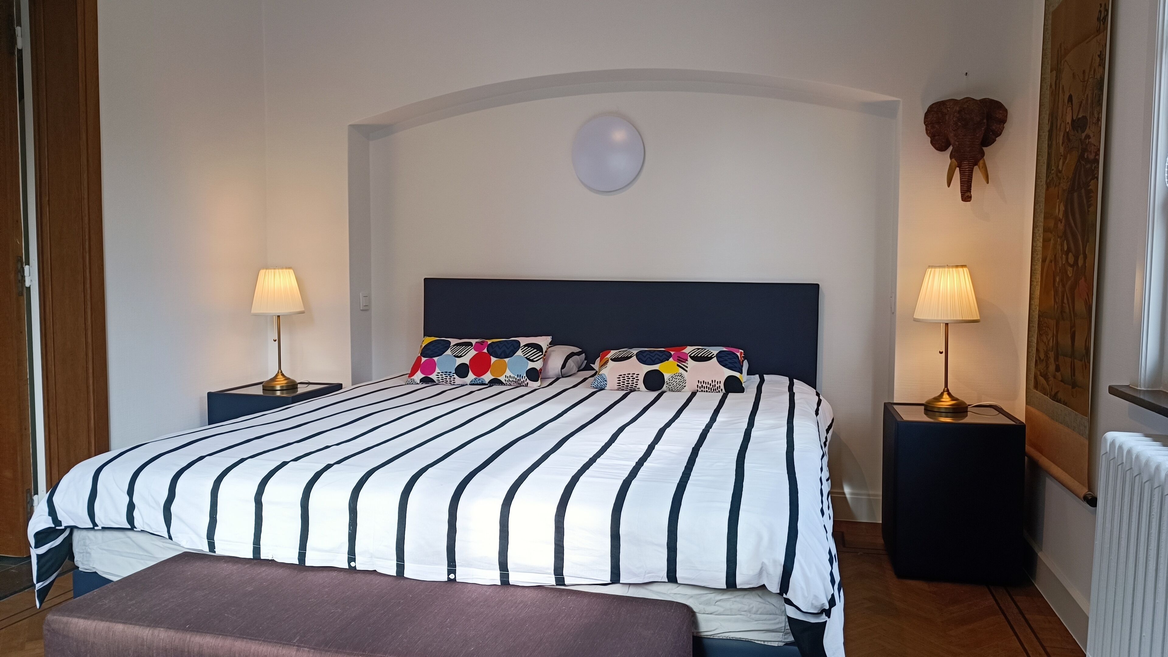 Ein Schlafzimmer mit einem großen Bett, zwei Nachttischen, einer Bank und einer Deckenleuchte.