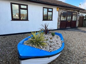 A house with a white exterior, a brown roof, and a blue boat-shaped planter filled with plants and pebbles in front.