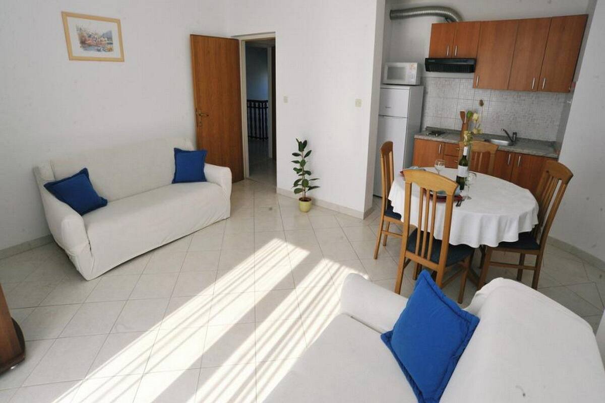 A living room with a white sofa, blue cushions, a dining table with chairs, a kitchen area with wooden cabinets, and a potted plant.