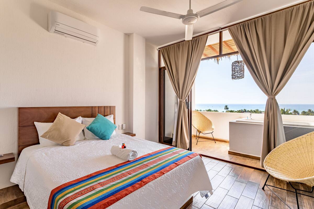 A hotel room with a bed, a ceiling fan, an air conditioning unit, a balcony with a view of the ocean, and a wicker chair.