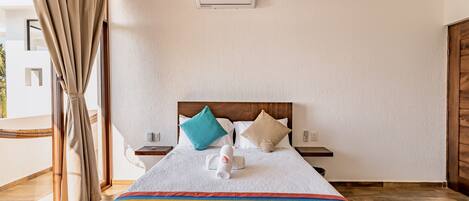 A hotel room with a large bed, a wooden headboard, two bedside tables, a nightstand, a window with curtains, and an air conditioning unit on the wall.