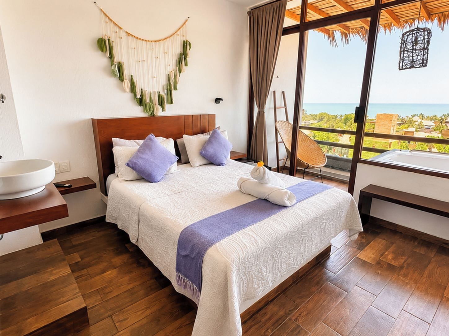 A hotel room with a large bed, a wooden headboard, a bedside table with a bowl, a view of the ocean, and a balcony with a wicker chair.