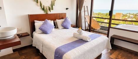 A hotel room with a large bed, a wooden headboard, a bedside table with a bowl, a view of the ocean, and a balcony with a wicker chair.