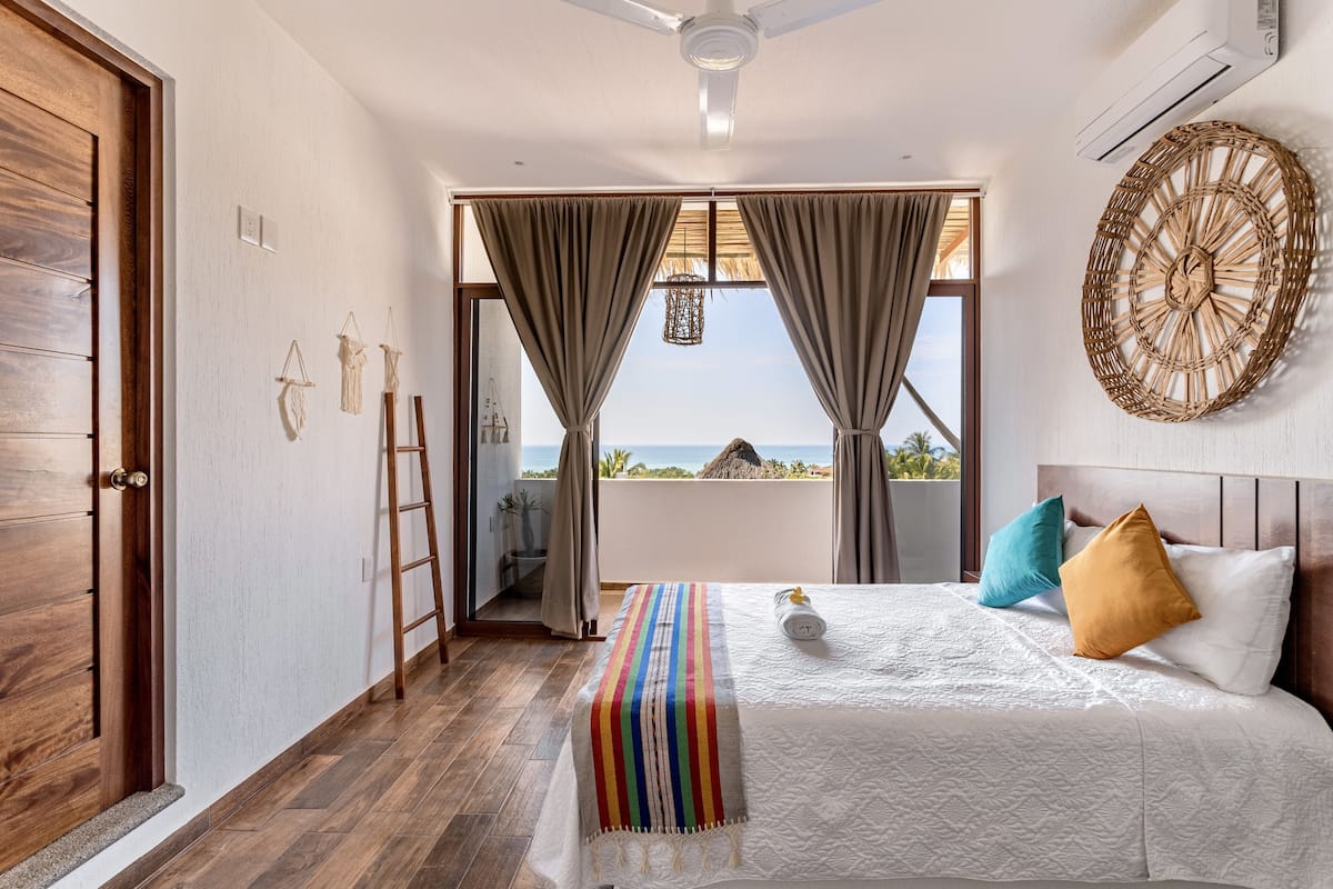 A bedroom with a large bed, wooden floor, a ceiling fan, a view of the sea, and a balcony with a thatched roof.