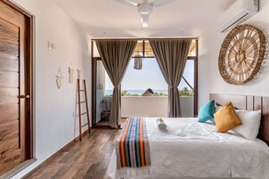 Un dormitorio con una cama grande, piso de madera, ventilador de techo, vista al mar y un balcón con techo de paja.