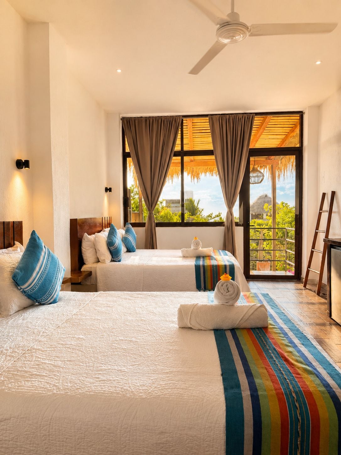 A hotel room with two beds, a ceiling fan, a large window with curtains, and a view of greenery outside.