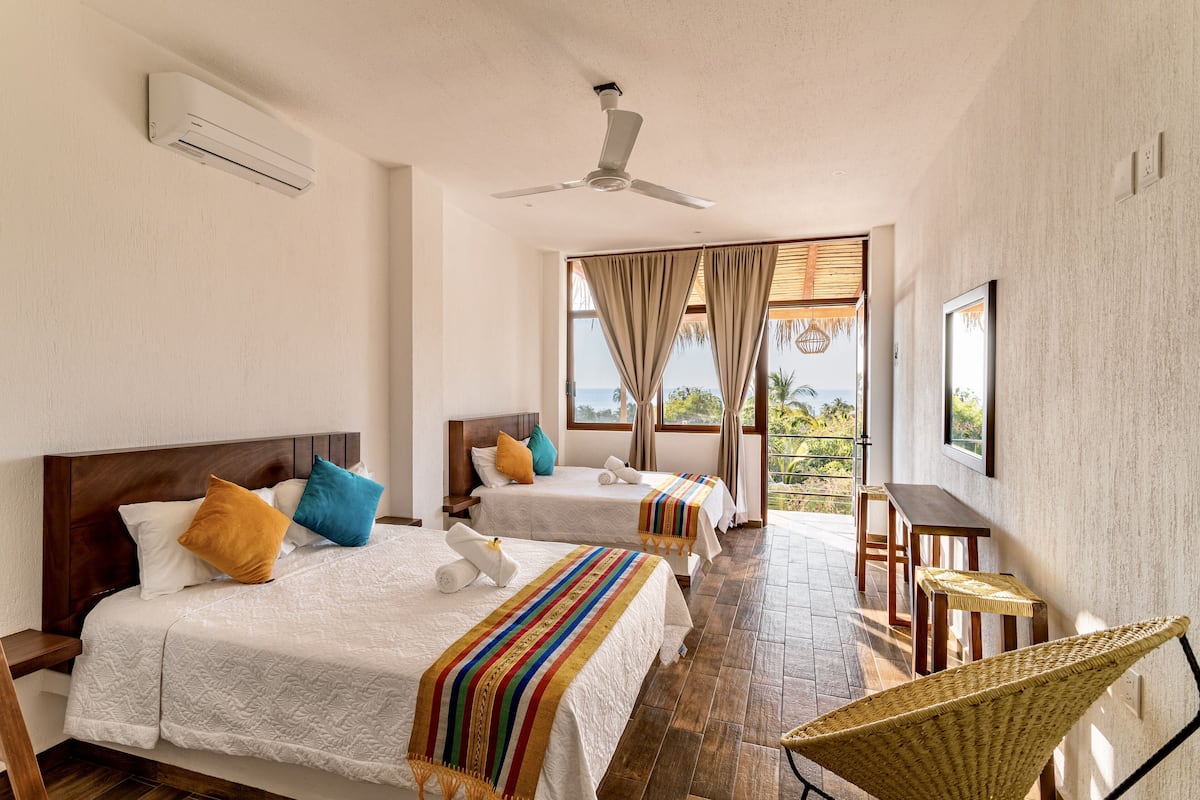 A hotel room with two beds, a ceiling fan, a dining table, and a wicker chair.