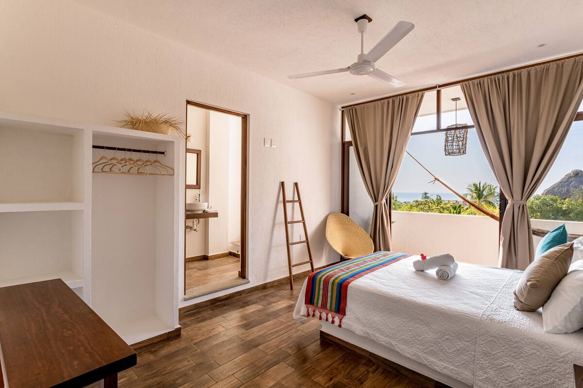 A bedroom with a bed, a wooden table, a wardrobe, a balcony with a view of the sea, and a ceiling fan.