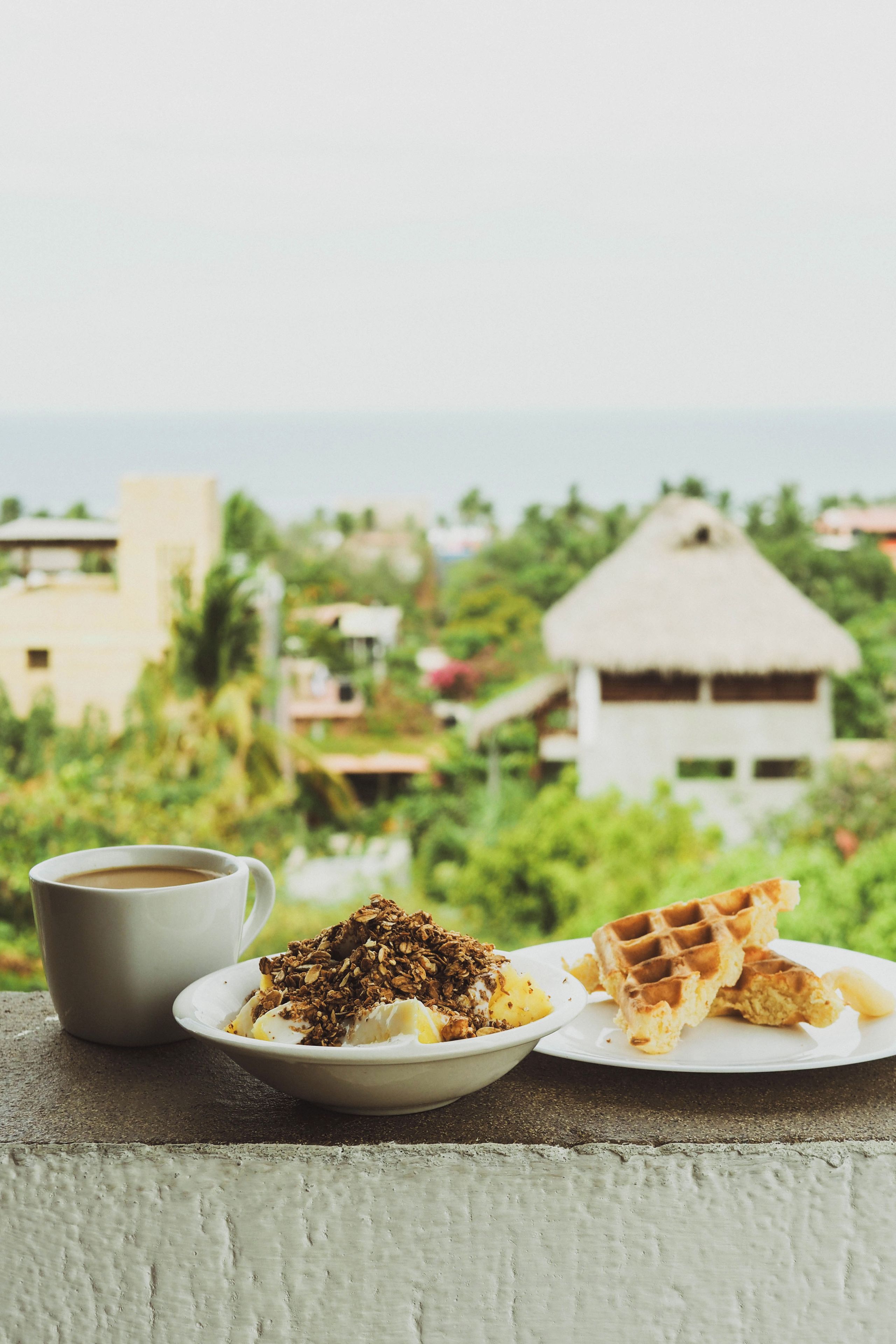 Un desayuno de yogur con granola y un gofre en un balcón con vistas a edificios y zonas verdes.